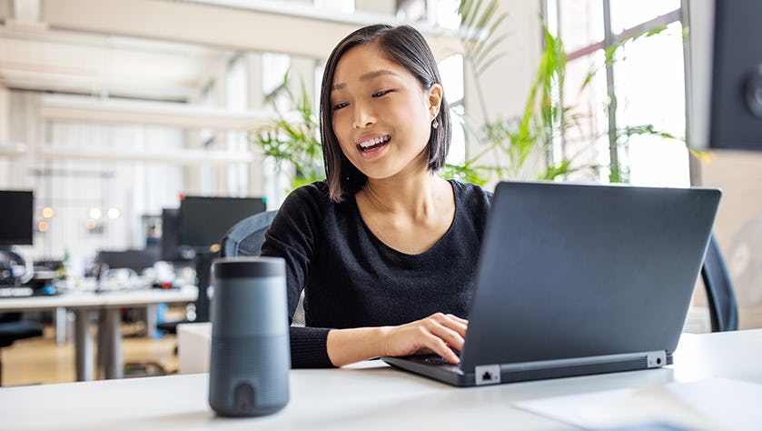 Asian businesswoman talking to virtual assistant at her desk. Female professional working on laptop and talking into a speaker.