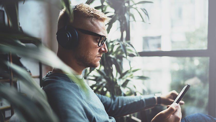 Young pensive man wearing eye glasses and casual clothes listening music in headphones on smartphone at modern place.Horizontal.Blurred background